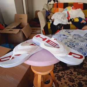 Vita flip-flops med röd text - Säljer ett par vita flip-flops med breda remmar och röd text på sulan. Skorna har en klassisk strandstil och är tillverkade i syntetmaterial. Perfekta för sommardagar och strandhäng.