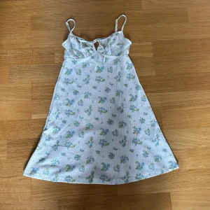 Blommig sommarklänning från Gina Tricot - Söt blommig klänning från Gina Tricot i vitt med blå och gröna blommor. Klänningen har smala axelband, knytning framtill och en lätt utsvängd passform. Perfekt för varma dagar!