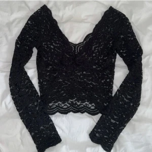 Svart spetsblus från Zara - Säljer en svart långärmad spetsblus från Zara med v-ringning och vågig kant. Blusen är croppad och har ett elegant spetsmönster över hela plagget. 