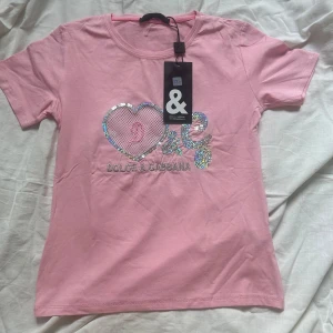 Rosa t-shirt från Dolce & Gabbana med paljettdekor - Snygg rosa t-shirt från Dolce & Gabbana med glittriga paljettdetaljer i form av hjärta och logga på bröstet. Klassisk rund halsringning och korta ärmar. Perfekt för dig som gillar statement-plagg med extra bling.