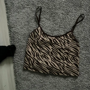Zebramönstrad croppad linnetopp - Säljer en croppad linnetopp med zebramönster i beige och brunt. Tunt ribbat material och smala axelband. Perfekt för sommaren och ger en trendig look.