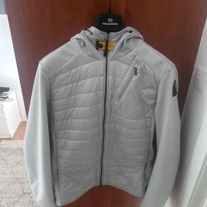  Parajumpers hybridjacka - Snygg ljusgrå hybridjacka från Parajumpers med quiltade partier framtill och på ryggen. Jackan har huva, dragkedja hela vägen, bröstficka med dragkedja och diskret logga på ärmen. Perfekt för lager-på-lager och stilren look. En månad gammal, ångrat köpet lite kommer ej till användning som jag trodde.