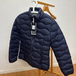 Mörkblå pufferjacka från Nimbus - Säljer en mörkblå pufferjacka från Nimbus med vågformat quiltat mönster. Jackan har hög krage, dragkedja framtill och två sidofickor. Perfekt för kyligare dagar och har en modern, sportig look.