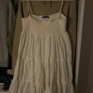 Vit sommarklänning med smock från Zara - Säljer en vit klänning från Zara med smock upptill och tunna axelband. Klänningen har en luftig, volangig kjol i flera lager och är perfekt för varma dagar. 💖