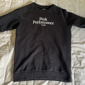 Svart sweatshirt från Peak Performance - Säljer en svart sweatshirt från Peak Performance med vit broderad logga på bröstet. Tröjan har rund halsringning, långa ärmar och ribbade muddar vid ärmslut och nederkant. Perfekt för en sportig och avslappnad stil.