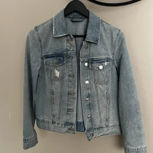 Klassisk ljusblå jeansjacka - Säljer en klassisk ljusblå jeansjacka med silverfärgade knappar och två bröstfickor. Jackan har lång ärm och en lätt tvättad look med diskreta slitningar. Perfekt att slänga över en hoodie eller t-shirt.