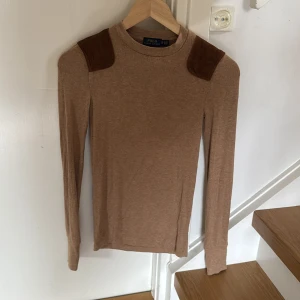 Beige långärmad tröja från Polo Ralph Lauren - Säljer en beige långärmad tröja från Polo Ralph Lauren med bruna axelpartier i mockaliknande material. Tröjan har en rund halsringning och är figurnära i modellen. Perfekt för en stilren och klassisk look.