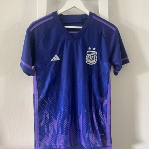 Argentina Adidas blå fotbollströja - Snygg Argentina fotbollströja från Adidas i blått och lila med korta ärmar. Tröjan har AFA-emblem och Adidas-logga på bröstet, samt lila detaljer på axlar och ärmslut. Tillverkad i lätt och ventilerande funktionsmaterial, perfekt för match eller träning.