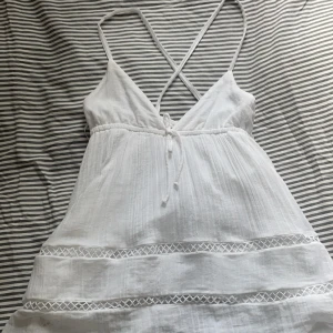 Vit somrig topp från Gina Tricot - Superfin vit topp från Gina Tricot med tunna axelband som korsas i ryggen. Klänningen har v-ringning, knyt framtill och dekorativa spetsdetaljer. Tillverkad i luftig bomull, perfekt för varma dagar.