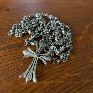 Chrome Hearts halsband - Säljer ett ikoniskt halsband från Chrome Hearts  med ett  korshänge och detaljerad kedja med blommönster. Kedjan har unika länkar och korset är graverat med märkets logga och årtal. Perfekt statement piece för dig som gillar edgy och exklusiv stil.