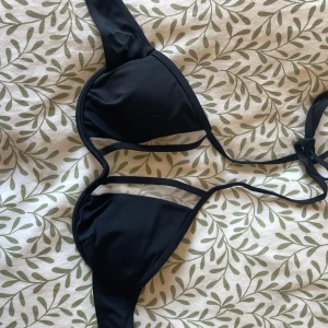 Svart bikini med knyt och metallspänne - Snygg svart bikini med trekantsformade kupor och knyt i ryggen. Toppen har ett stilrent metallspänne bak och smala band. Perfekt för stranden eller poolen, enkel att matcha med andra plagg. Klassisk och tidlös design.