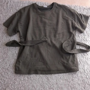 Knyttopp  - Olivgrön t-shirt från Jacqueline de Yong i mjuk bomull. Modellen har korta ärmar, rund halsringning och coola knytband i midjan för en snygg twist. Perfekt till jeans eller kjol för en chill look. Tyvärr har sömmen gått av där bak men det syns inte när man knyter ☺️