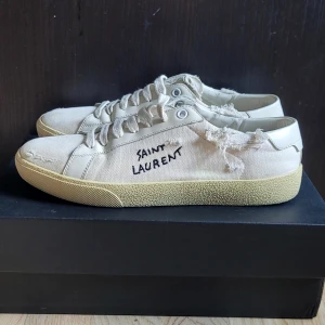 Saint Laurent SL/06 i beige canvas - Saint Laurent sneakers i beige canvas med cool sliten look och handskriven logga på sidan. Låg modell med snörning och robust beige sula. Perfekta för dig som gillar en avslappnad stil och exklusiva detaljer. Skorna samt og medföljer. Nypris 7000kr mitt pris 2499kr🔥 Kontakta mig om frågor och fler bilder, priset är inte fast så kom med prisförslag.