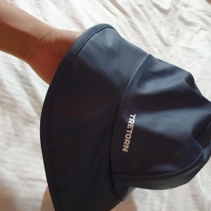 Marinblå regnhatt från Tretorn - Snygg marinblå regnhatt från Tretorn med bred brätte och justerbar knäppning under hakan. Tillverkad i vattentätt syntetmaterial med mjukt grått foder på insidan. Perfekt för regniga dagar och håller dig torr med stil.