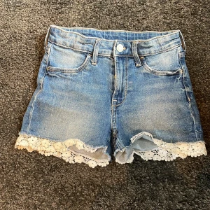 Blå jeansshorts med spetskant strl. 136 - Snygga blå jeansshorts med ljus tvätt och vit blommig spetskant längst ner. Klassisk femficksmodell med bälteshällor och knappstängning. Perfekta för sommarens varma väder☀️💐