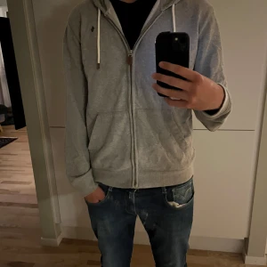 Grå hoodie från Morris med dragkedja - Säljer en grå hoodie från Morris med vit snörning i huvan och dragkedja framtill. Tröjan har två fickor och en liten broderad logga på bröstet. Perfekt för chill dagar eller när du vill ha en enkel och stilren look. 