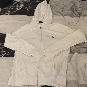 Vit hoodie från Ralph Lauren - Vit hoodie från Polo Ralph Lauren med dragkedja och huva med snörning. Klassisk broderad logga i marinblått på bröstet och två fickor framtill. Säljer eftersom den inte längre passar mig.