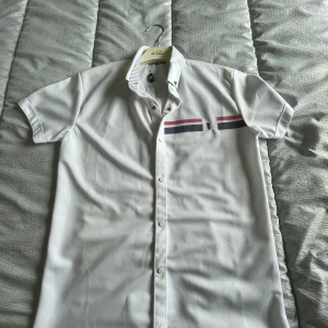 Vit kortärmad skjorta U.S. Polo Assn. - Vit kortärmad skjorta från U.S. Polo Assn. med slim fit-passform. Skjortan har knappar framtill, detaljerade röda och mörkblå ränder på bröstet samt broderad logga. Kragen har ett diskret svartvitt mönster på insidan. Perfekt för en clean och sportig stil.några flickor på skjortan.man kan få bort de med ett medel 