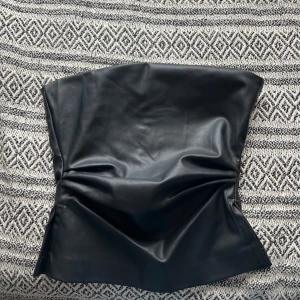 Svart skinn tubtopp - Säljer en svart tubtopp i skinnimitation. Toppen är axelbandslös och har en stilren, minimalistisk look som sitter tight runt kroppen. Perfekt för dig som vill ha en edgy vibe till outfiten.