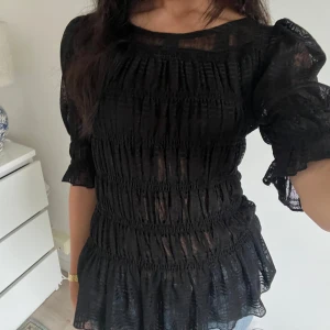 Svart blus med volang och puffärm - Säljer en svart blus med transparent tyg, smockad överdel och volang nertill. Blusen har puffiga trekvartsärmar med volangkant och en luftig, flowy känsla. Perfekt att styla med jeans eller kjol för en trendig look.