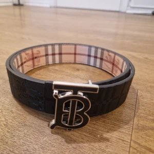 Svart bälte  - Stilren svart bälte med rutigt mönster i beige, rött och svart på baksiden, som du också kan använda som framsida. Storlek 110 cm, och priset går att diskutera