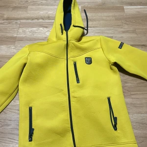Gul vindjacka från Claswin XXL - Gul vindjacka från Claswin i storlek XXL. Jackan har huva med dragsko, svarta dragkedjor på fickor och framtill samt snygga logodetaljer på bröstet och ärmen. Perfekt för dig som vill sticka ut med färg och sportig stil.