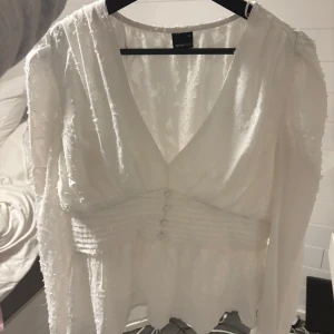 Vit v-ringad blus från Gina Tricot - Supersnygg vit blus från Gina Tricot med v-ringning och markerad midja med knappar framtill. Lätt textur i tyget och långa ärmar ger en trendig look. Perfekt att styla med jeans eller kjol för en chic vibe. Liten i storleken