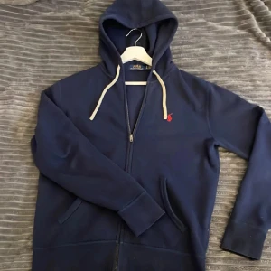 Mörkblå hoodie från Polo Ralph Lauren - Säljer en mörkblå hoodie från Polo Ralph Lauren med dragkedja och vit snörning i huvan. Tröjan har det klassiska röda Polo-logot på bröstet, kängurufickor och ribbade muddar. Perfekt för chill dagar och snygg till jeans eller joggers.