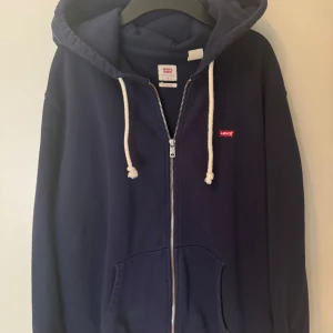 Mörkblå hoodie från Levi's med dragkedja - Mörkblå hoodie från Levi's i bomull med vit snörning i huvan och dragkedja framtill. Klassisk passform med känguruficka och liten röd Levi's-logga på bröstet. Perfekt för chill dagar och enkel att matcha med jeans.