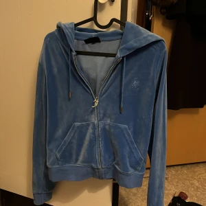 Blå velour hoodie Juicy Couture S - Säljer en ikonisk blå velour hoodie från Juicy Couture i storlek S. Tröjan har huva, dragkedja framtill, snörning vid huvan och klassiska kängurufickor. Perfekt för en chill och trendig look, med broderad logga på bröstet.