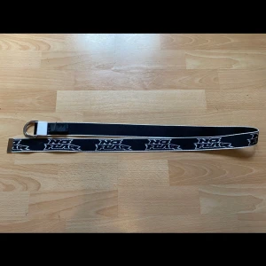 Svart No Fear x H&M tygbälte - Svart tygbälte från No Fear x H&M med vit logga längs hela bältet. Bältet har dubbla silverfärgade D-ringar som spänne och vita kanter. Perfekt accessoar för att ge din outfit en streetwear-vibe.