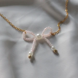 Guldhalsband med pärlrosett - Sött halsband med tunn guldfärgad kedja och en rosett av ljusrosa och vita pärlor, dekorerad med små guldfärgade kulor. Perfekt för dig som gillar gulliga detaljer och pärlor.