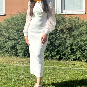 Vit långklänning med spetsärmar - Elegant vit långklänning med v-ringning och draperad överdel. Klänningen har genomskinliga spetsärmar med broderade blommor och pärldetaljer på axlarna. Materialet är glansigt och känns lyxigt, perfekt för speciella tillfällen.