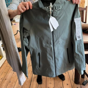 Grön regnjacka från MaxMara - Snygg grön regnjacka från MaxMara med minimalistisk design och hel dragkedja framtill. Jackan har två praktiska fickor med dragkedja och hög krage. Perfekt för regniga dagar och enkel att matcha med olika outfits.