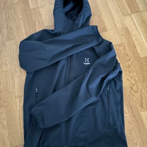 Svart vindjacka från Haglöfs, stl L - Svart vindjacka från Haglöfs i storlek L. Jackan har huva, hel dragkedja framtill och två sidofickor med dragkedja. Tillverkad i lätt och slitstark polyester, perfekt för blåsiga dagar. Snygg minimalistisk design med broderad Haglöfs-logga på bröstet.
