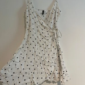 Vit omlottklänning med svarta prickar - Vit omlottklänning från H&M devided med svarta prickar. Klänningen har smala axelband, knyt i sidan och en flowy, kort passform. 
