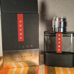 Prada Luna Rossa Carbon EdT 50ml - Prada Luna Rossa Carbon Eau de Toilette 50ml med elegant svart och silverfärgad glasflaska. Flaskan har en röd vertikal detalj med Prada-loggan och en robust metallkork. Doften är modern och maskulin, perfekt för dig som gillar exklusiva parfymer. Knappt använd. 