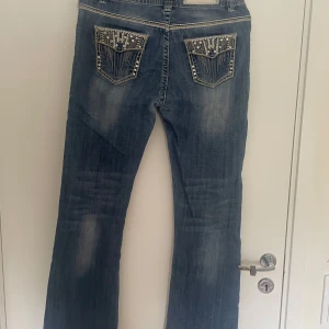 Rock & Roll blå jeans med detaljer - Snygga blå jeans från Rock & Roll med coola broderade och glittriga detaljer på fickorna och längs sidorna. Jeansen har kontrastsömmar, normal passform och klassisk femficksdesign. Perfekta för dig som gillar statement-plagg och unika detaljer. Säljs för att dem inte passade som ja tänkte.litet hål som är igensatt som inte syns skriv för bild. mått= längd:102cm. innerbenslängd=80cm. midjemått:38cm rakt över