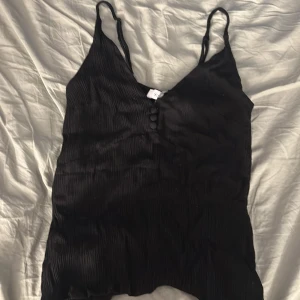 Svart ribbad linnetopp Stylein XS - Supersnygg svart linnetopp från Stylein i storlek XS. Toppen har tunna axelband, v-ringning och ribbad struktur. Dekorativa knappar framtill ger en extra touch. Det är ett litet hål vid v-ringningen som enkelt kan sys