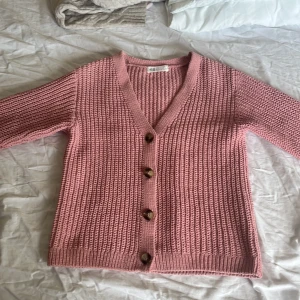 Rosa stickad kofta från H&M - Superfin rosa stickad kofta från H&M med v-ringning och fyra stora knappar framtill. Koftan har en chunky ribbad struktur, perfekt att slänga på sig över en t-shirt eller ett linne. Enkel och snygg att styla till jeans eller kjol.
