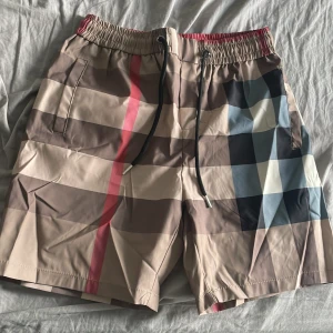 Rutig badshorts med snörning - Snygga badshorts med klassiskt rutigt mönster i beige, brunt, svart, blått och en röd rand. Shortsen har elastisk midja med svart snörning och sidofickor. Perfekta för stranden eller poolen, med en modern och trendig look.