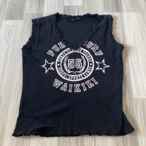 Svart ärmlös topp med tryck - Svart ärmlös topp med v-ringning och kanter med volanger. Har ett sportigt tryck