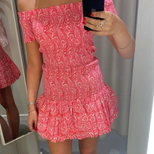 Rosa offshoulder kortklänning - Superfin rosa kortklänning med vitt mönster och offshoulder-modell. Klänningen har smockad överdel och volangig kjol, perfekt för sommarens alla tillfällen. Snygg och bekväm passform som framhäver axlarna.