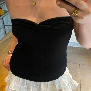 Svart bandeau tubtopp Divided H&M - Svart bandeau tubtopp från Divided by H&M i stretchig bomullsmix. Toppen är axelbandslös med twistad detalj framtill och sitter tight mot kroppen. Perfekt för en trendig och enkel look.