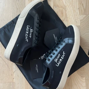 Svarta Saint Laurent sneakers - Snygga svarta sneakers från Saint Laurent med vit logga broderad på sidan. Skorna har svarta snören, vit platt sula och detaljer i skinn och textil. Perfekta för dig som gillar stilrena och trendiga skor med exklusiv känsla.
