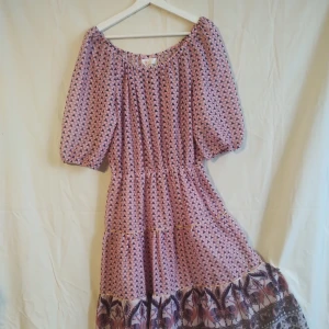 Långklänning i lila mönster - Långklänning med lila och rosa geometriskt mönster, puffiga korta ärmar och volangkant nertill med extra mönsterdetaljer. Klänningen har en lös passform och är tillverkad i ett lätt, glittrigt syntetmaterial. Perfekt för dig som gillar boho-vibes och unika prints.