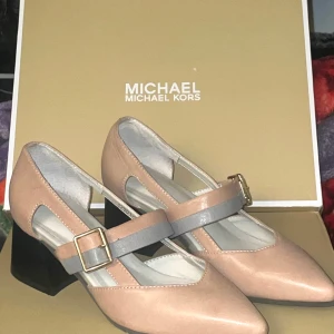 Beiga pumps med blockklack från Michael Kors - Stilrena pumps i beige skinn från Michael Kors med spetsig tå och bred svart blockklack. Skorna har två remmar över foten, en beige och en grå, båda med guldfärgat spänne. Perfekta för dig som gillar klassisk men modern stil.