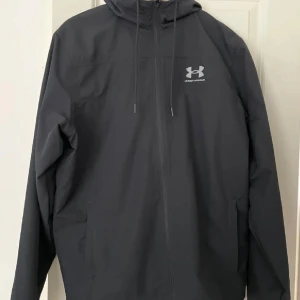Svart vindjacka från Under Armour - Snygg svart vindjacka från Under Armour med huva och dragsko. Jackan har dragkedja framtill, två sidofickor och en stilren logga på bröstet. Perfekt för dig som gillar sportig och clean stil.