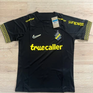 AIK svart matchtröja Nike M - AIK:s officiella svarta matchtröja från Nike med gula detaljer och klubbmärke på bröstet. Truecaller-logga framtill, SVEA-logga bak och gula mönster på ärmsluten. Tillverkad i lätt och ventilerande polyester, perfekt för fotbollsträning eller match.