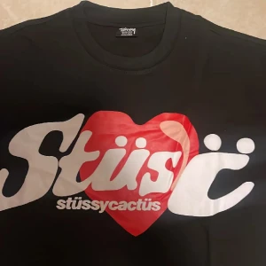 Svart Stüssy x Cactus t-shirt med hjärta - Svart t-shirt från Stüssy med stort vitt tryck och rött hjärta på bröstet där det står 'stüssycactus'. Klassisk passform med rund hals och korta ärmar. Perfekt för dig som gillar streetwear och vill sticka ut med en unik design.